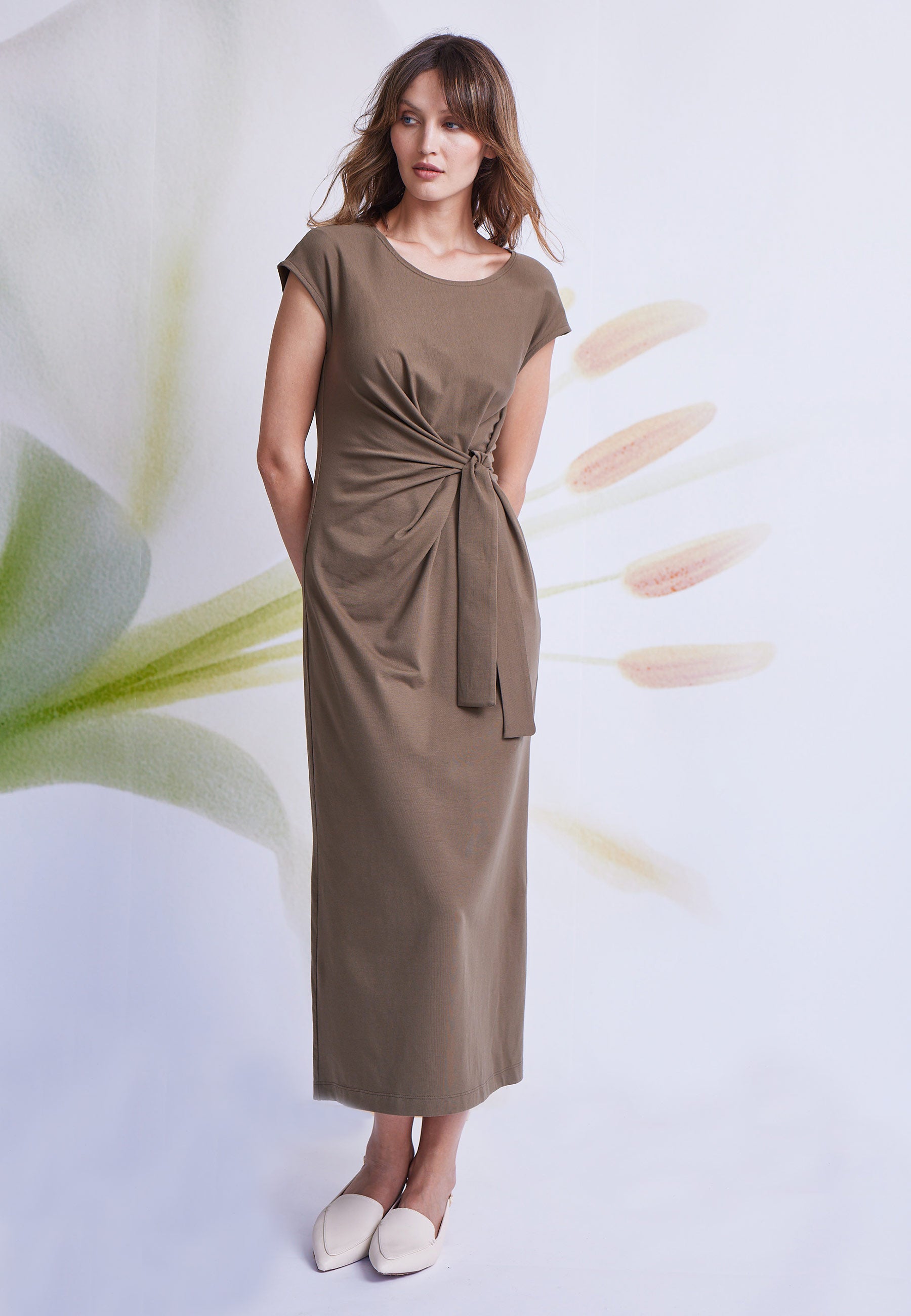 Kleid, Modell Marly in warmem Braun mit kurzem Arm und dekorativem Knotendetail, stehend vor floralem Hintergrund