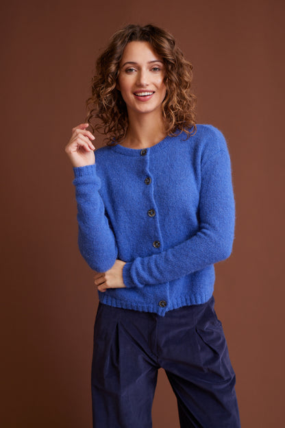 Frau trägt blaue, weich gestrickte Jacke mit Knöpfen, Modell Nikola, kombiniert zu dunkelblauer Hose vor braunem Hintergrund