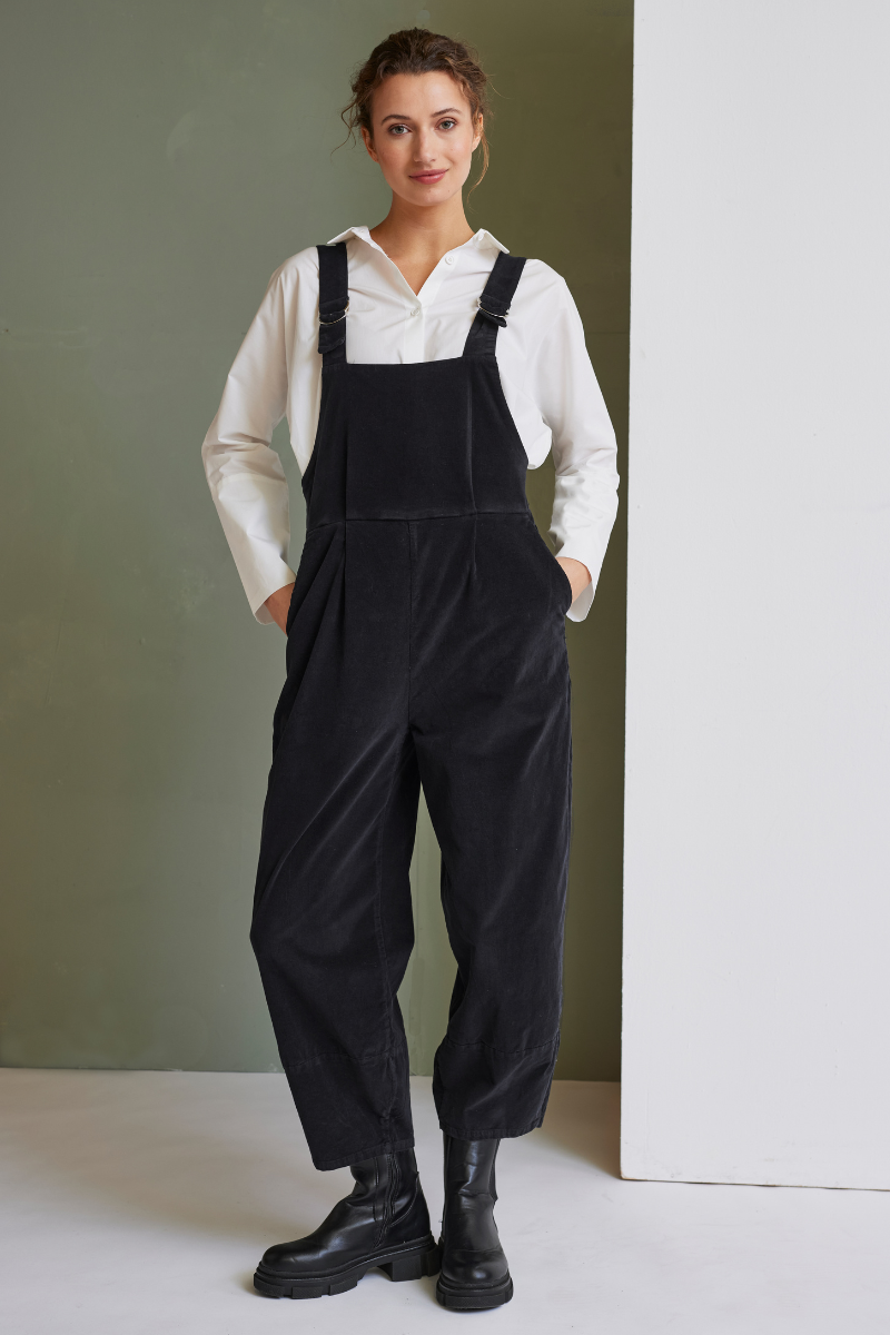 Frau trägt schwarze Trägerhose, Modell Emmet, mit weißem Hemd und schwarzen Boots vor grün-weißem Hintergrund